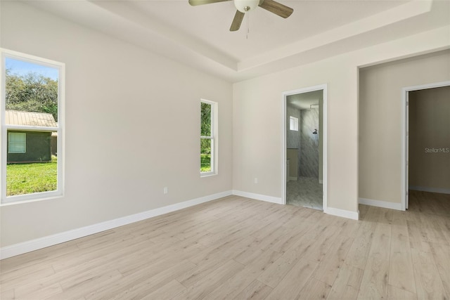 unfurnished bedroom featuring light wood finished floors, baseboards, a tray ceiling, ensuite bathroom, and a ceiling fan