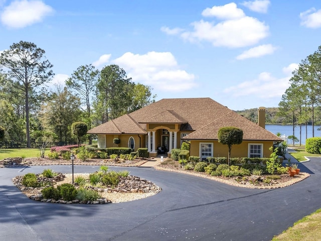 view of front facade with stucco siding, a water view, curved driveway, fence, and a shingled roof