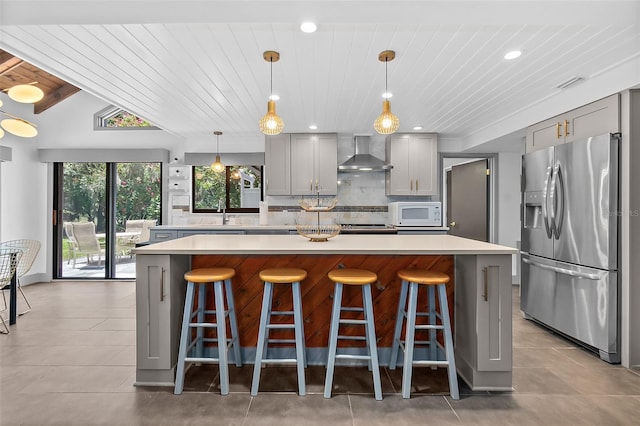 kitchen with wall chimney range hood, white microwave, backsplash, stainless steel refrigerator with ice dispenser, and gray cabinetry