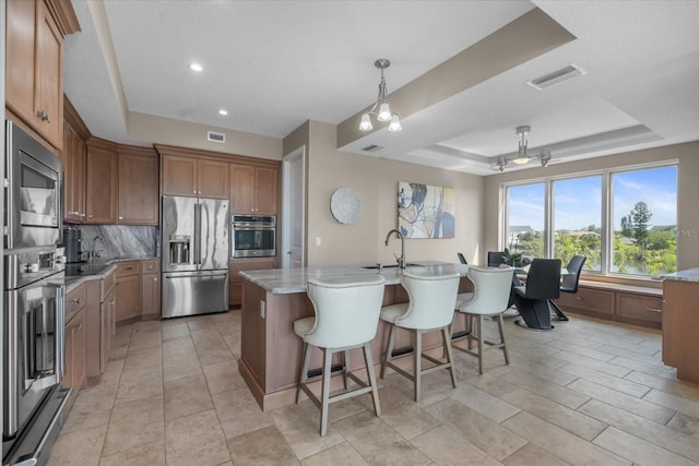 2590 Palm Ave, Flagler Beach, FL 32136 - kitchen with stainless steel appliances, a raised ceiling, decorative backsplash, a center island with sink, and brown cabinetry