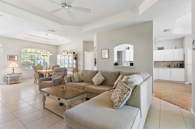 living room featuring ceiling fan with notable chandelier, baseboards, light tile patterned floors, a tray ceiling, and visible vents