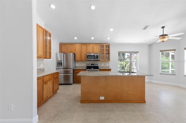 kitchen featuring visible vents, glass insert cabinets, an island with sink, tasteful backsplash, and stainless steel appliances