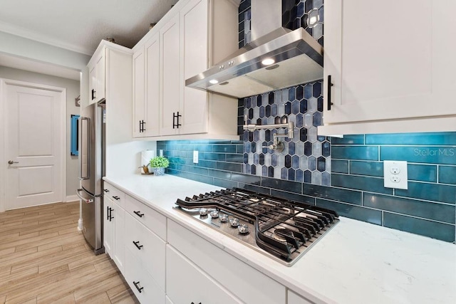 kitchen with wall chimney range hood, stainless steel appliances, white cabinetry, light wood-style flooring, and decorative backsplash