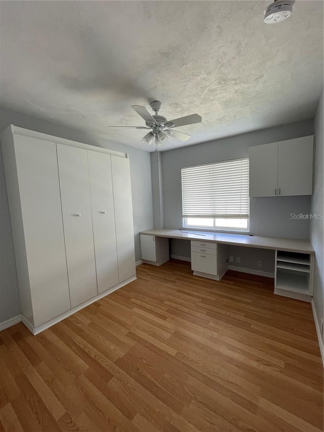 unfurnished office featuring built in study area, light wood-style flooring, ceiling fan, and baseboards