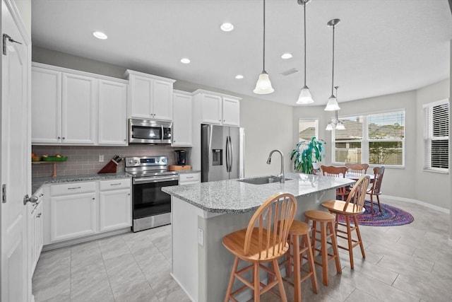 kitchen with stainless steel appliances, a sink, backsplash, light stone countertops, and a center island with sink