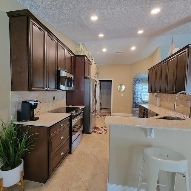 kitchen with stainless steel appliances, a sink, arched walkways, dark brown cabinetry, and a textured ceiling