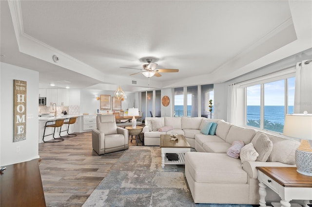 living room featuring a tray ceiling, wood finished floors, a ceiling fan, crown molding, and a water view