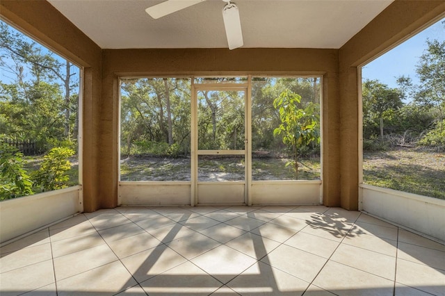 unfurnished sunroom with tile patterned floors, ceiling fan, plenty of natural light, and view of wooded area