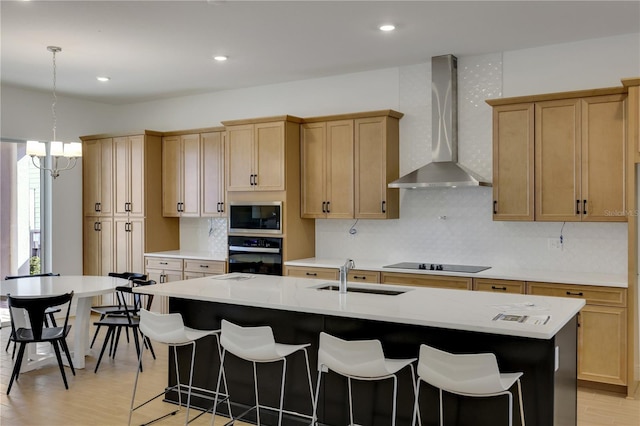 kitchen with pendant lighting, wall chimney range hood, sink, black appliances, and an island with sink