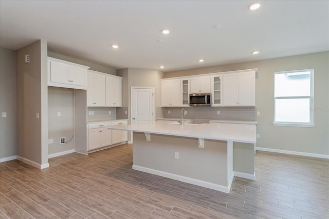 kitchen featuring light countertops, stainless steel microwave, decorative backsplash, white cabinets, and an island with sink