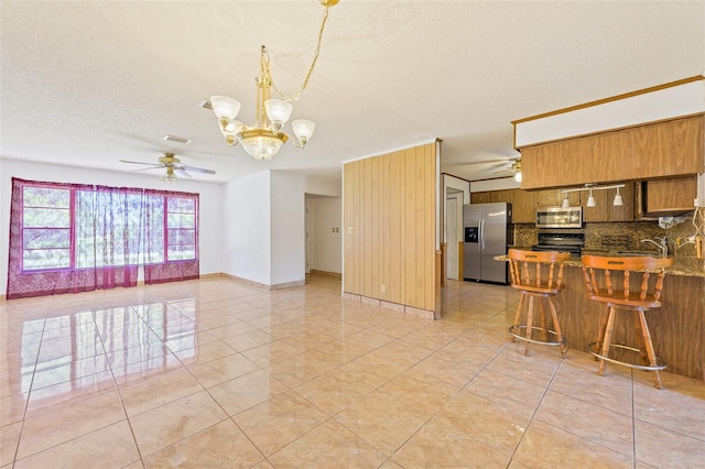 kitchen with appliances with stainless steel finishes, light tile floors, backsplash, ceiling fan with notable chandelier, and a kitchen breakfast bar