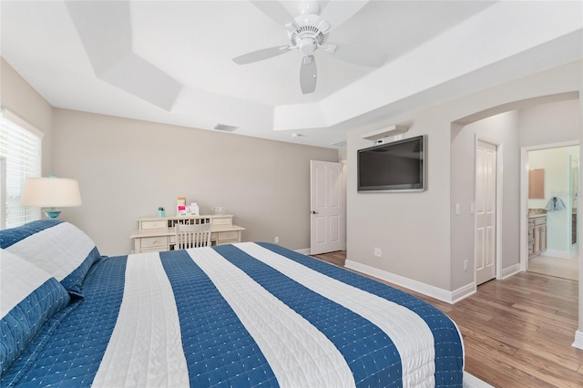 bedroom with ceiling fan, wood-type flooring, a tray ceiling, and ensuite bath