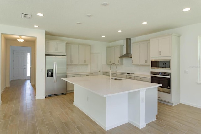 kitchen featuring appliances with stainless steel finishes, sink, wall chimney range hood, white cabinetry, and an island with sink