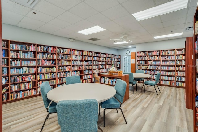 living area featuring light wood-type flooring, a drop ceiling, ceiling fan, and wall of books