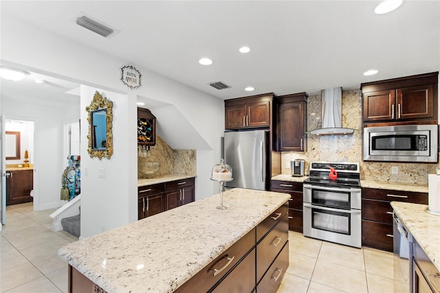 kitchen featuring wall chimney range hood, light tile patterned flooring, appliances with stainless steel finishes, a center island, and light stone counters