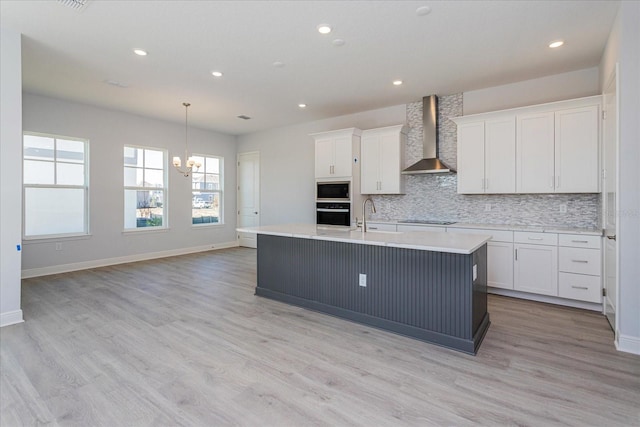 kitchen with white cabinetry, an island with sink, backsplash, black appliances, and wall chimney range hood