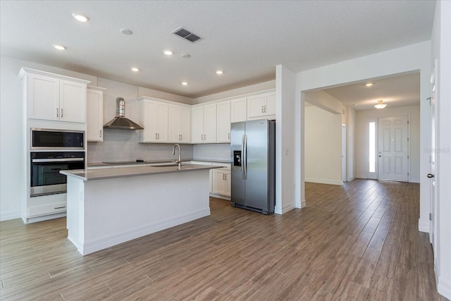 kitchen with wall chimney exhaust hood, appliances with stainless steel finishes, white cabinets, and an island with sink