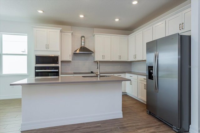 kitchen featuring appliances with stainless steel finishes, tasteful backsplash, wall chimney range hood, an island with sink, and white cabinets