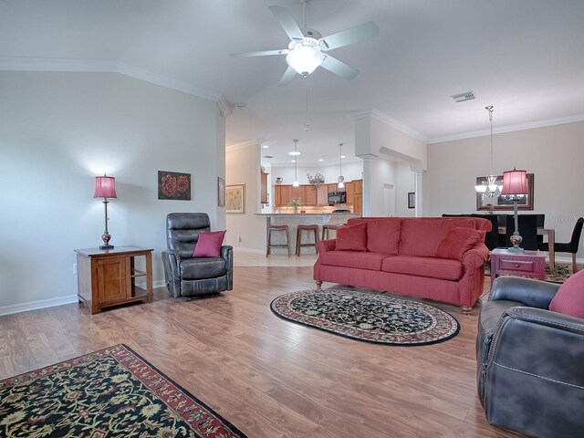 living area with decorative columns, crown molding, visible vents, light wood-style floors, and ceiling fan with notable chandelier