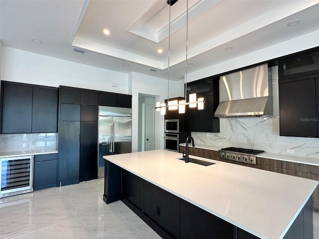 kitchen featuring pendant lighting, sink, a large island with sink, built in appliances, and wall chimney range hood