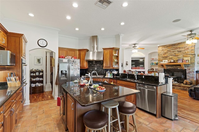 kitchen featuring ceiling fan, stainless steel appliances, wall chimney range hood, and a fireplace