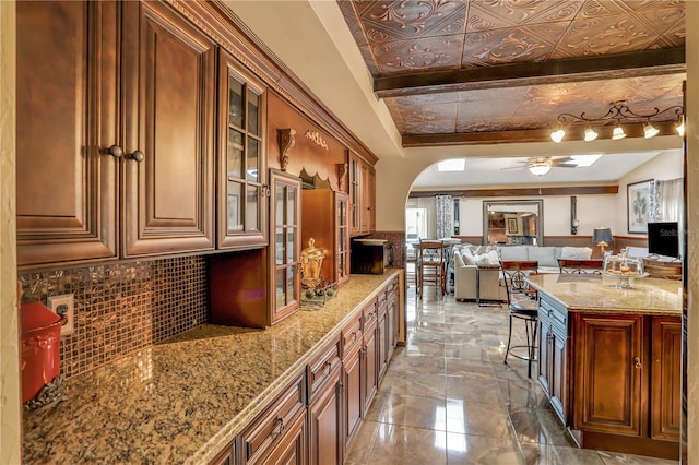 kitchen with decorative backsplash, a kitchen breakfast bar, light stone counters, ceiling fan, and beam ceiling