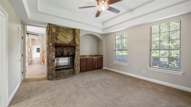 unfurnished living room featuring ceiling fan, a stone fireplace, light carpet, and a tray ceiling