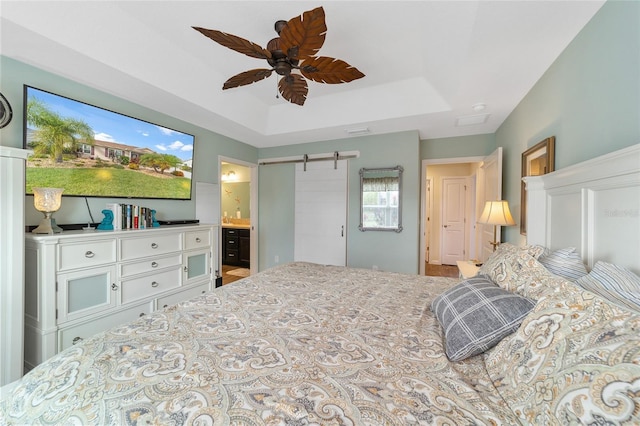 bedroom featuring ceiling fan, a barn door, a tray ceiling, and ensuite bath