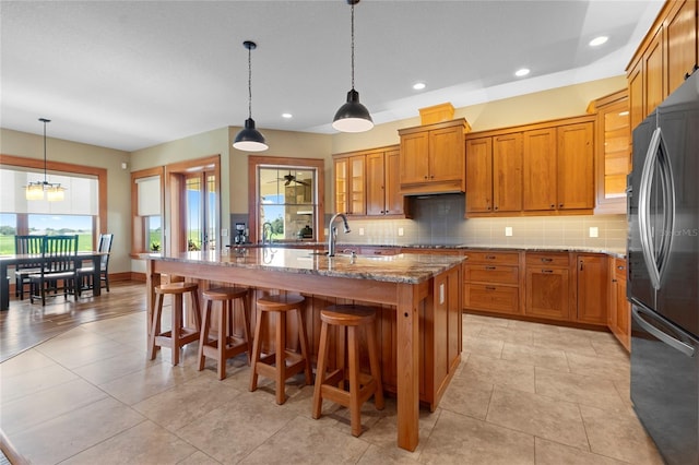 kitchen with stainless steel refrigerator, light stone counters, a kitchen island with sink, and hanging light fixtures