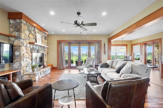 living room featuring a stone fireplace, ceiling fan with notable chandelier, light hardwood / wood-style floors, and french doors