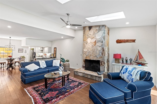 living room featuring a stone fireplace, ceiling fan with notable chandelier, wood-type flooring, and a skylight