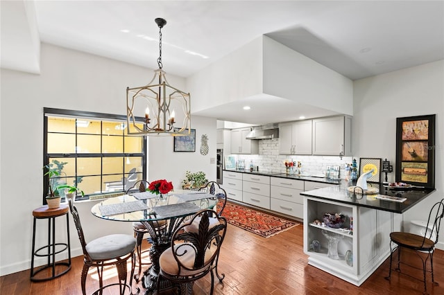 kitchen featuring kitchen peninsula, backsplash, a chandelier, dark wood-type flooring, and wall chimney exhaust hood