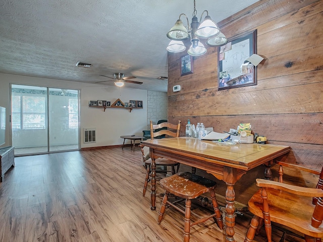 dining area with ceiling fan with notable chandelier, a textured ceiling, wooden walls, and hardwood / wood-style flooring