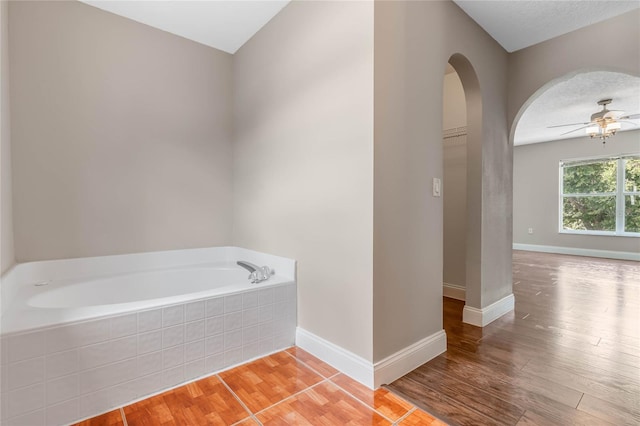 bathroom with a textured ceiling, tiled tub, wood-type flooring, and ceiling fan