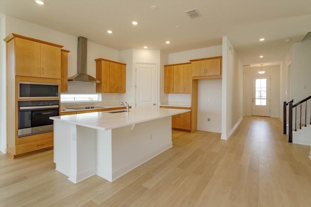 kitchen with built in microwave, sink, stainless steel oven, a center island with sink, and wall chimney range hood