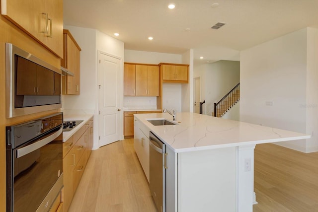 kitchen with sink, a kitchen island with sink, stainless steel appliances, light stone countertops, and light hardwood / wood-style flooring