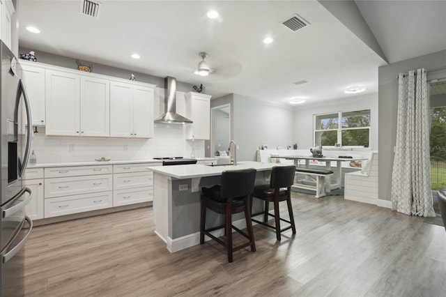 kitchen featuring an island with sink, decorative backsplash, wall chimney exhaust hood, white cabinets, and light hardwood / wood-style floors