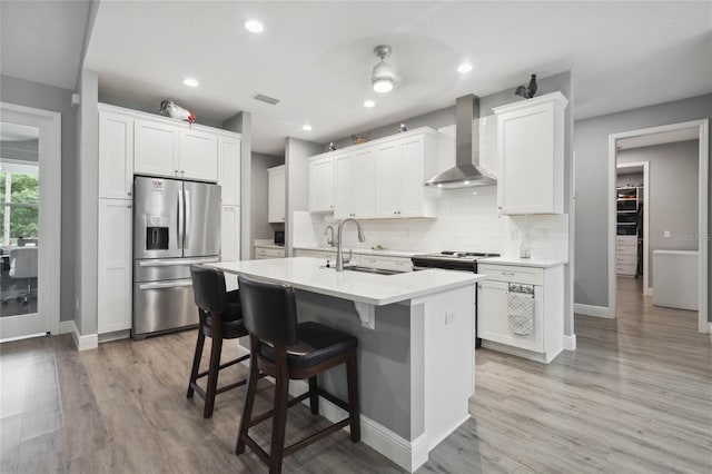 kitchen featuring a center island with sink, stainless steel appliances, light wood-type flooring, and wall chimney range hood