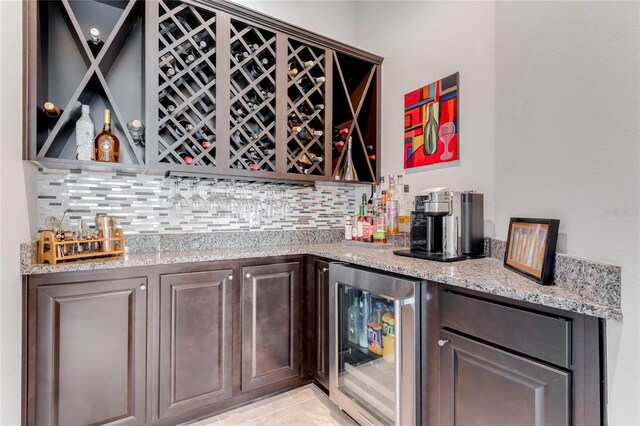 bar with light tile patterned flooring, beverage cooler, decorative backsplash, dark brown cabinetry, and light stone countertops