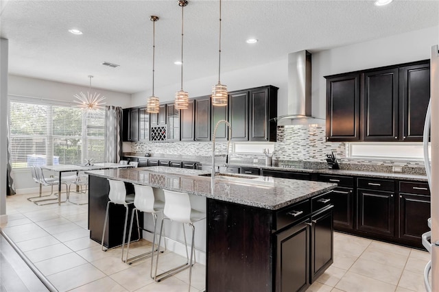 kitchen with backsplash, pendant lighting, wall chimney exhaust hood, light stone countertops, and a center island with sink