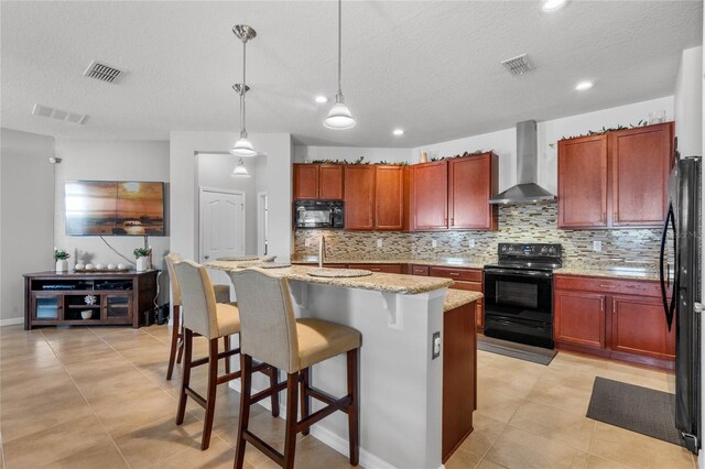 kitchen with light tile patterned floors, a kitchen island with sink, backsplash, black appliances, and wall chimney exhaust hood