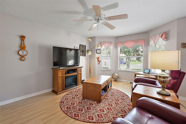 living area featuring light wood finished floors, a glass covered fireplace, ceiling fan, a textured ceiling, and baseboards