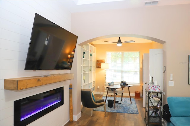 living area featuring arched walkways, visible vents, a ceiling fan, a glass covered fireplace, and wood finished floors