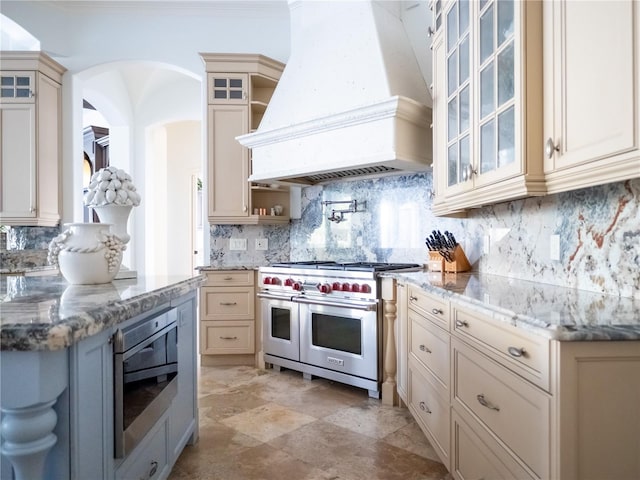 kitchen with light stone countertops, stainless steel appliances, cream cabinets, decorative backsplash, and custom exhaust hood