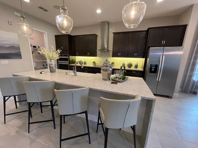 kitchen featuring wall chimney range hood, appliances with stainless steel finishes, an island with sink, and a sink