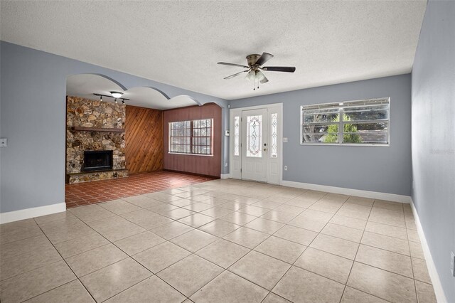 unfurnished living room with a textured ceiling, ceiling fan, wooden walls, light tile patterned floors, and a stone fireplace