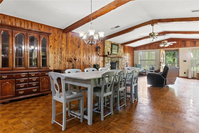 dining area with wooden walls, ceiling fan with notable chandelier, a stone fireplace, dark parquet floors, and vaulted ceiling with beams