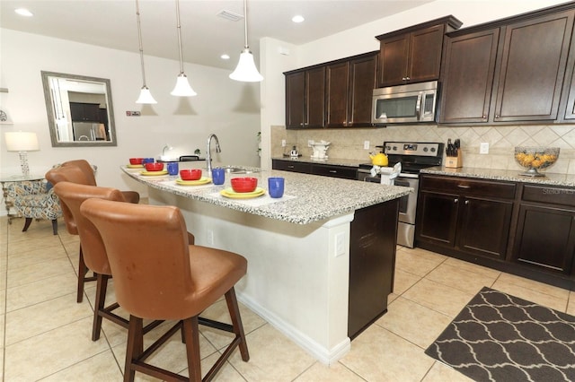 kitchen featuring light stone countertops, hanging light fixtures, appliances with stainless steel finishes, a breakfast bar area, and a center island with sink