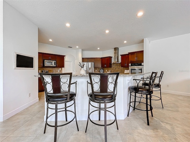 kitchen with a breakfast bar area, appliances with stainless steel finishes, wall chimney exhaust hood, and a kitchen island with sink