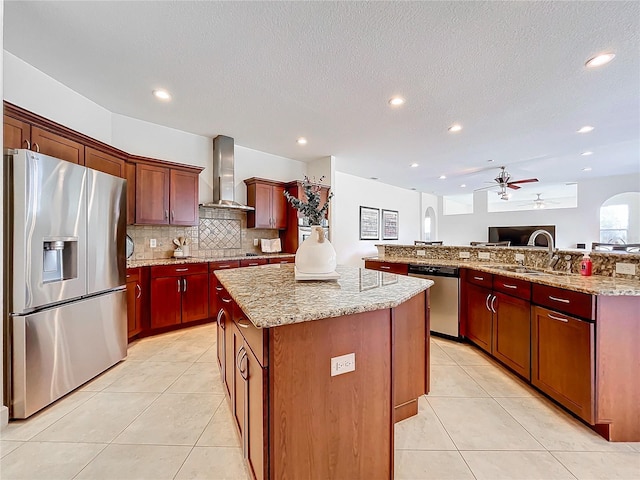 kitchen featuring a kitchen island, wall chimney range hood, stainless steel appliances, light stone countertops, and ceiling fan